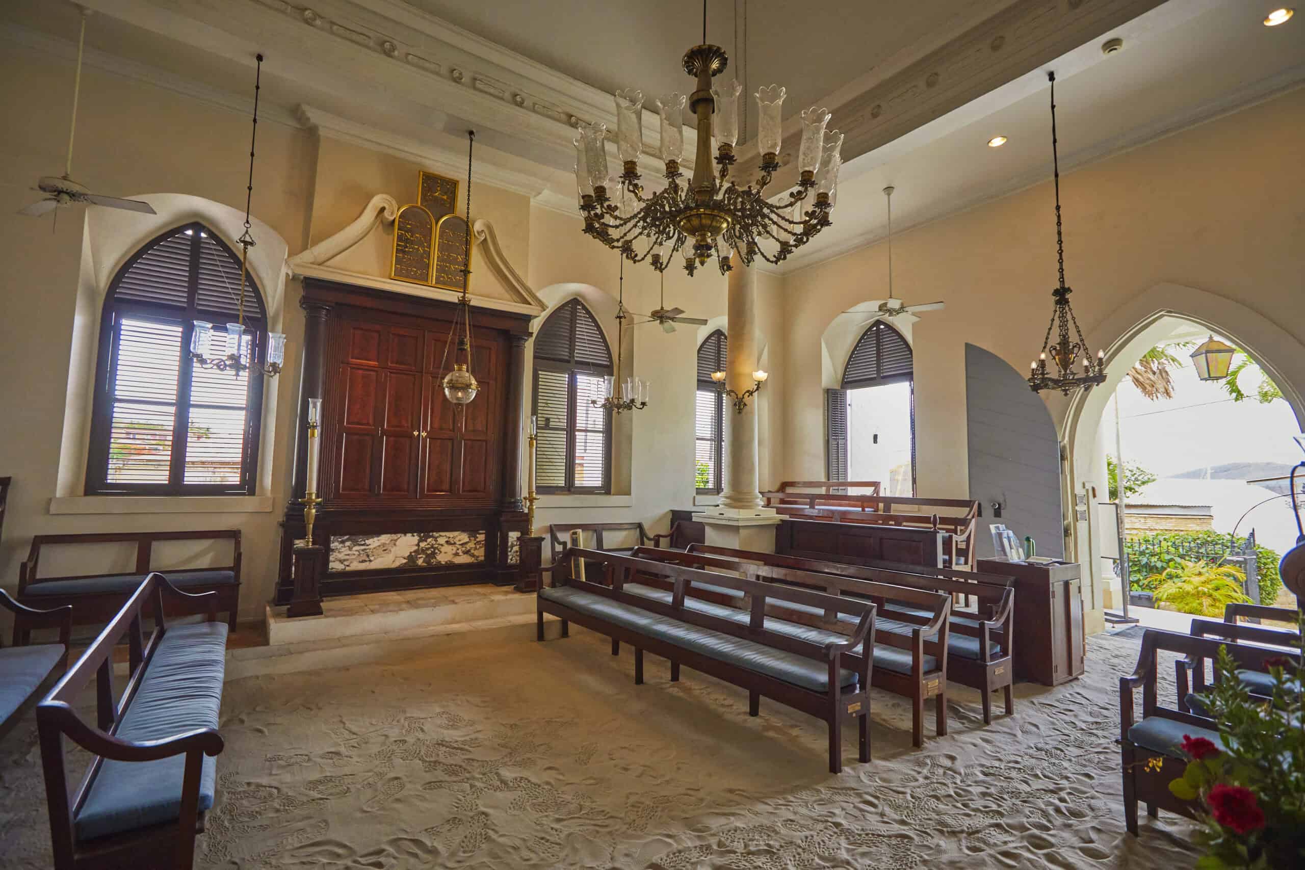 Interior of a synagogue with wooden benches and a large chandelier. The floor is covered with sand, and arched windows allow natural light in.