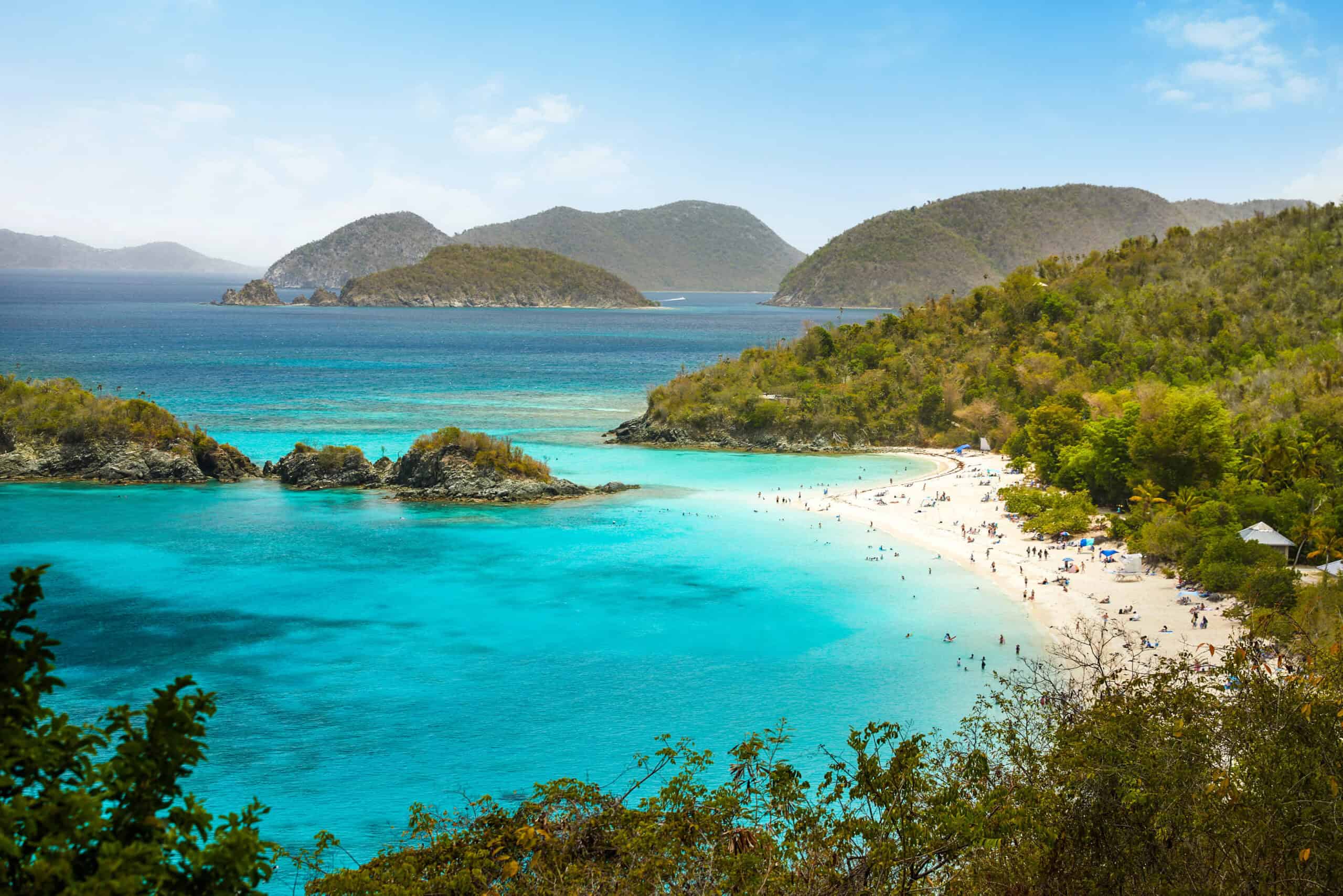 Overhead view of a tropical beach with turquoise water, surrounded by green hills and small islands under a blue sky. People are visible on the sandy shoreline of Trunk Bay Beach on St. John, USVI.