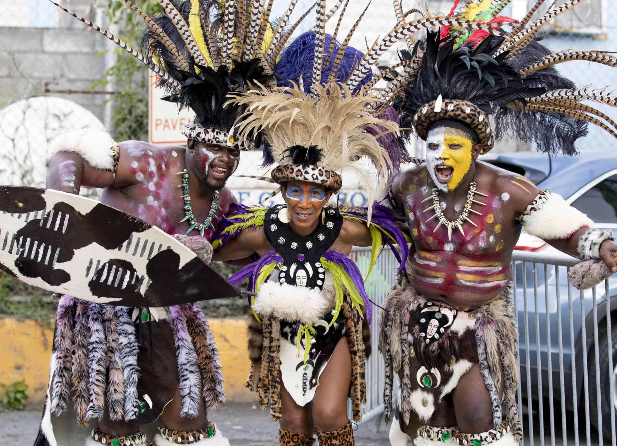 Three people in vibrant, feathered costumes with body paint pose energetically outdoors at a USVI Carnival celebration.