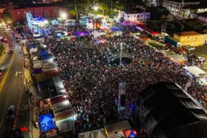Aerial view of a large crowd at an outdoor festival with colorful lights, stages, and booths, surrounded by streets with cars.