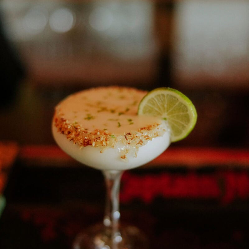A creamy cocktail in a coupe glass with a toasted coconut rim, garnished with a lime wheel, sits on a dimly lit bar at the Lovango Rum Bar in St. John, U.S. Virgin Islands.