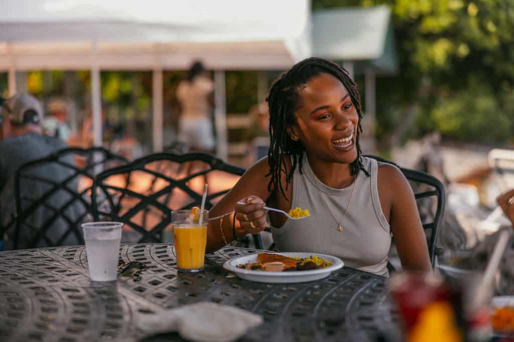 A woman sits at an outdoor table at Miss Lucy's on St. John smiling and eating a meal with a fork; a glass of orange juice and a cup of water are beside her.