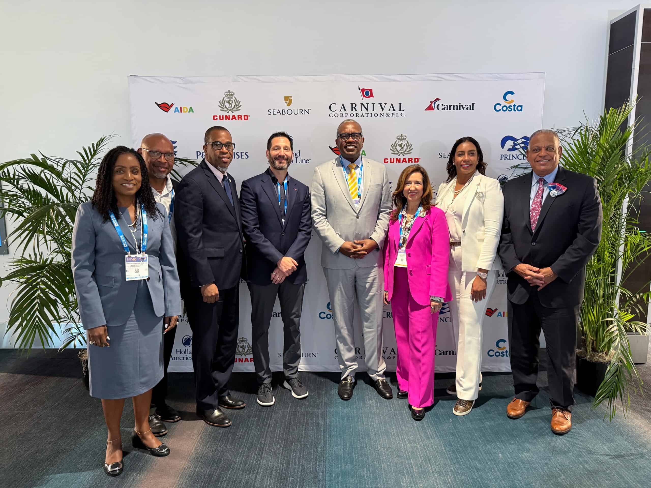 Eight professionally dressed individuals stand in a row, posing for a group photo in front of a backdrop with Carnival Corporation and cruise line logos.