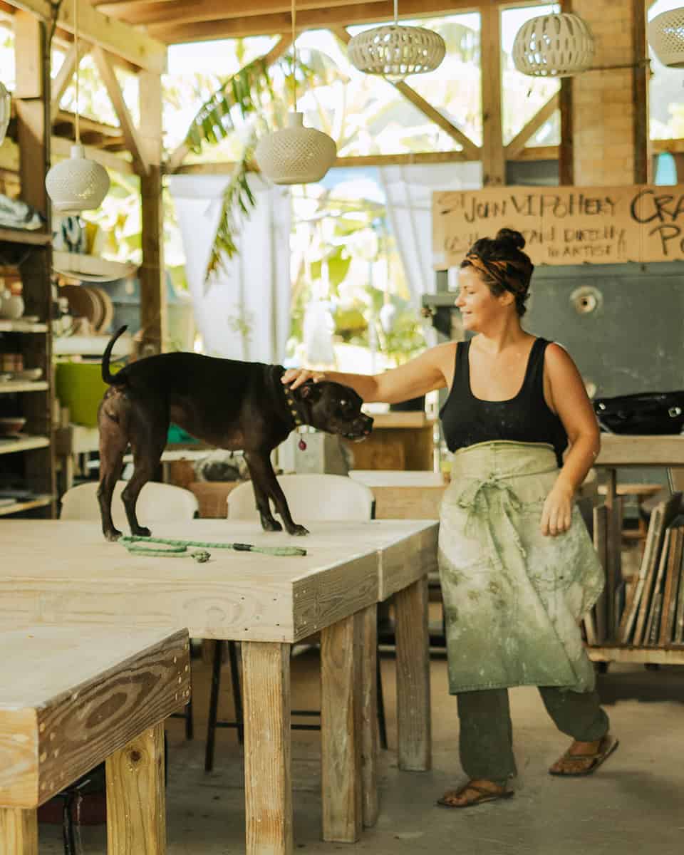 Woman in a workshop pets a dog standing on a wooden table. Hanging lights and various supplies are visible in the background.