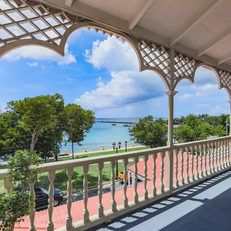 View from a porch with decorative arches, overlooking a coastal scene with trees, a bridge over the water, and a partly cloudy sky.