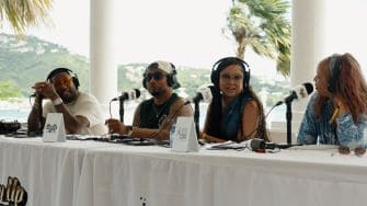 Four people wearing headphones sit at a long table with microphones, participating in a panel discussion outdoors with palm trees in the background.