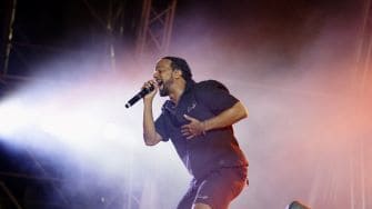 A man wearing dark clothing sings passionately into a microphone on a stage with dramatic lighting and visible stage structures in the background.