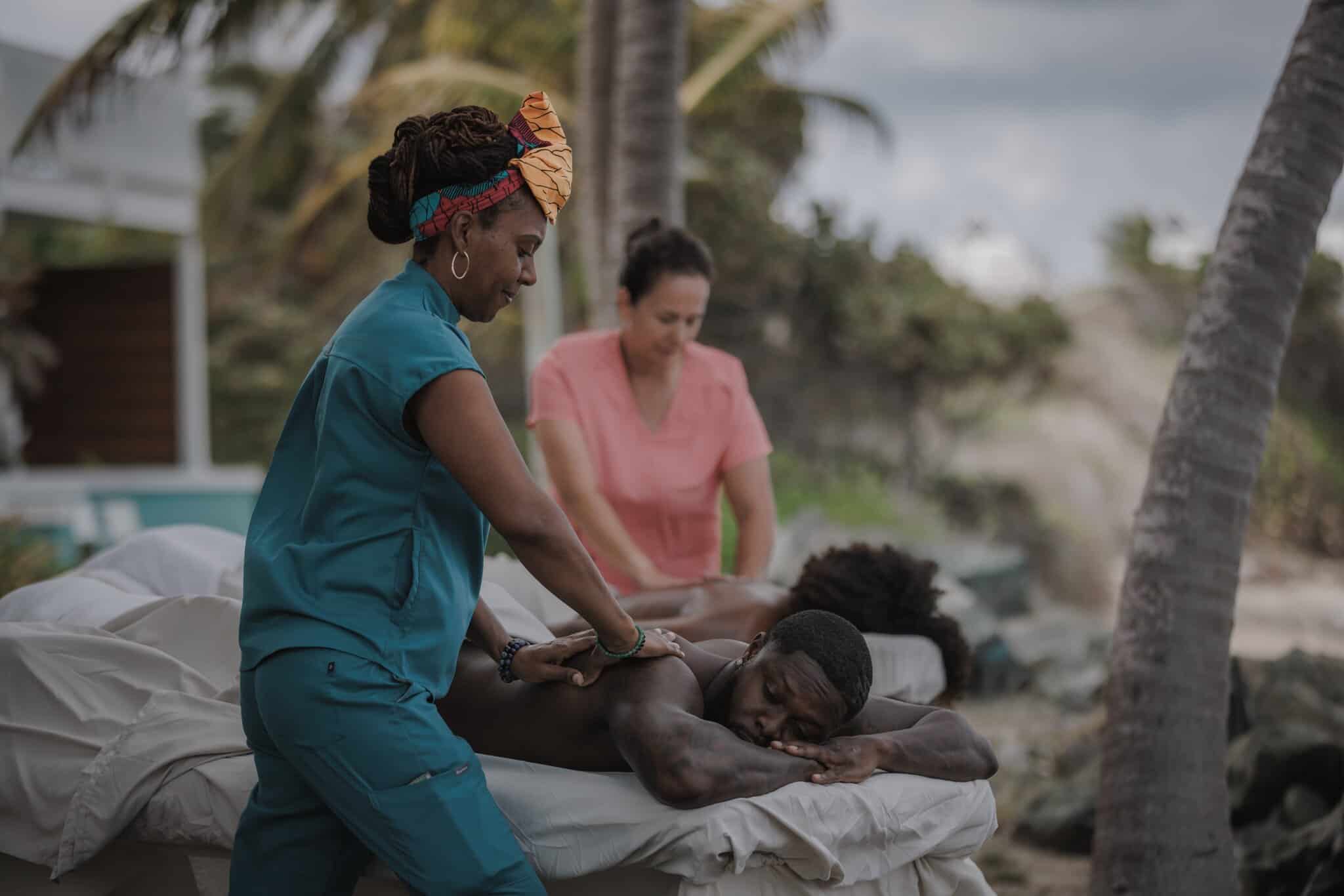 Two massage therapists give outdoor massages to clients on tables under palm trees, with a tropical landscape in the background.