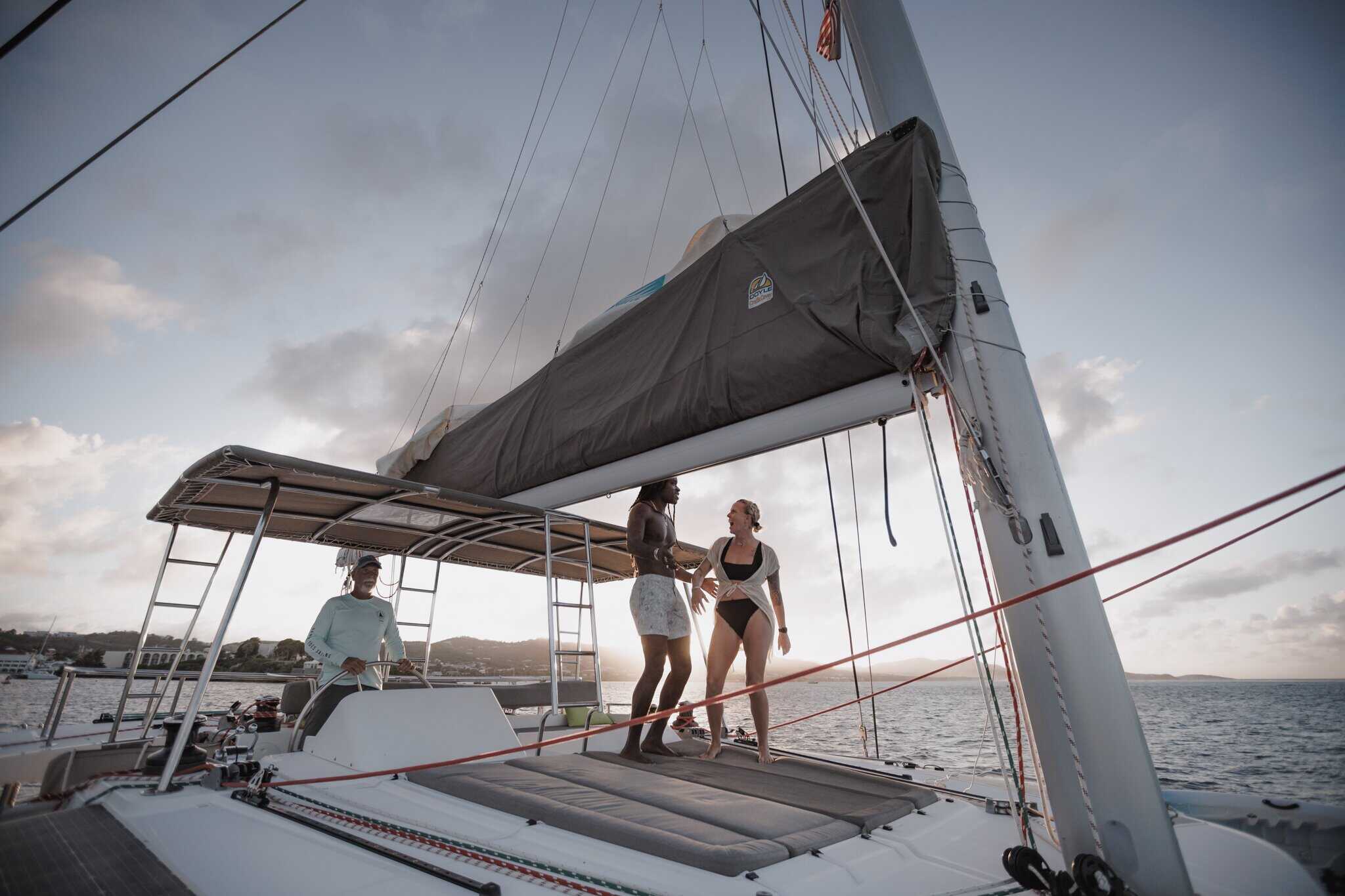Three people are on a sailboat at sunset; two are standing and talking near the sails, while one is sitting under the canopy. The sea and coastline are visible in the background.