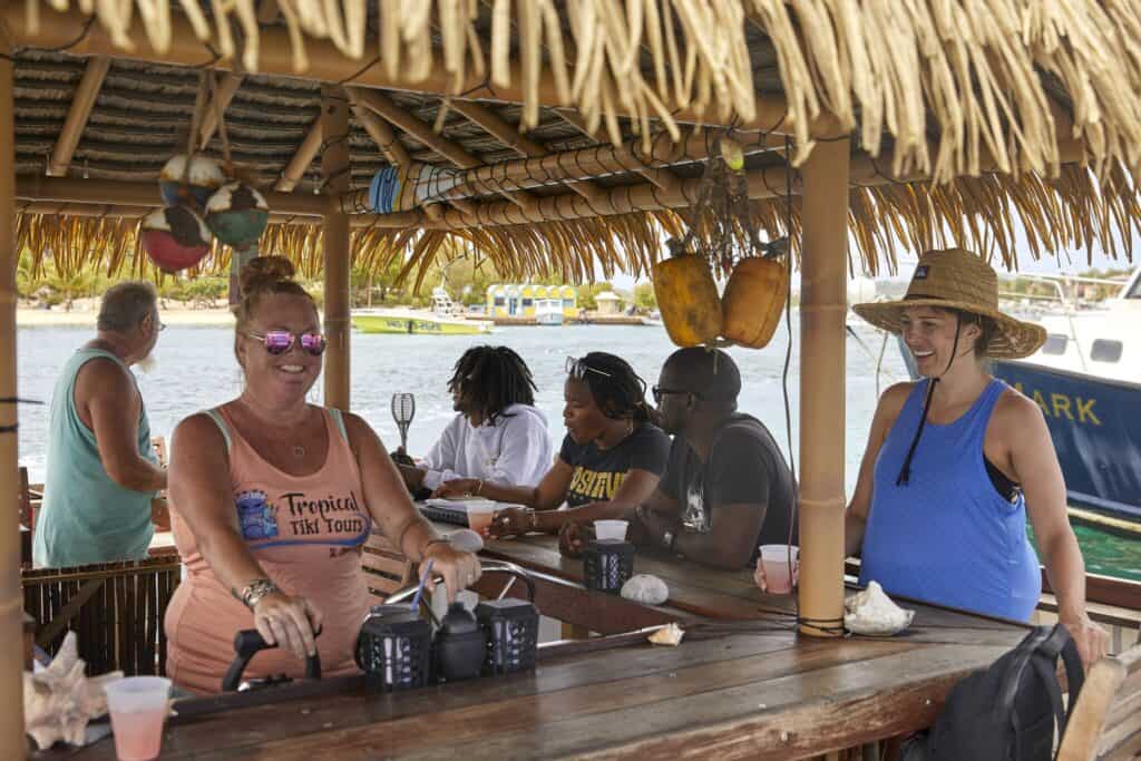A bartender smiles as she prepares a drink at a tiki bar on St. Croix. A person is laughing as they sit in one of the chairs to order.