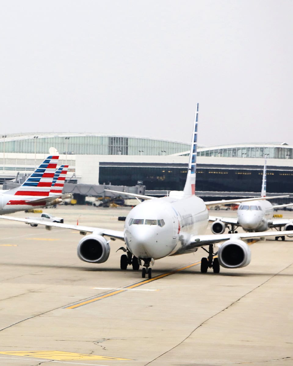 Commercial airplanes taxi on the runway at an airport, with terminal buildings visible in the background under a cloudy sky.