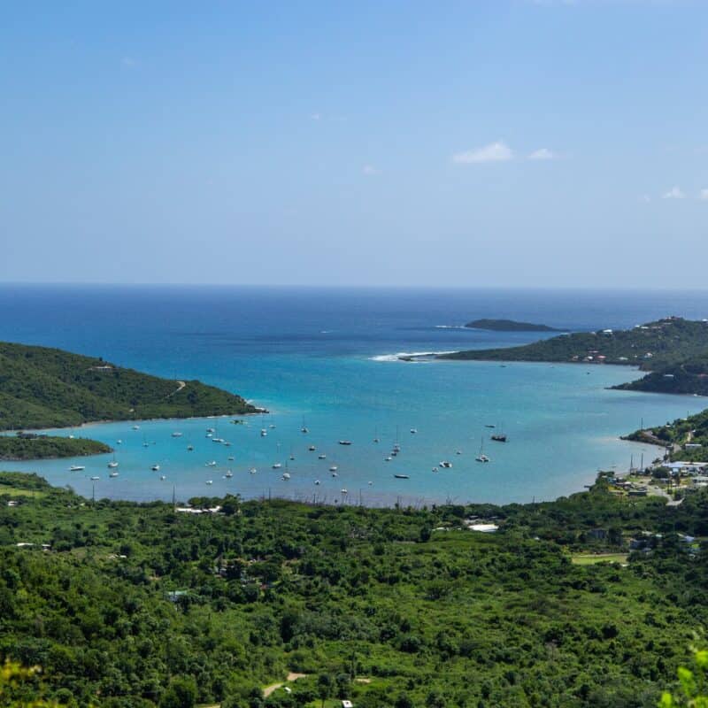 A coastal bay with turquoise water, scattered boats, green hills, and small buildings under a clear blue sky.