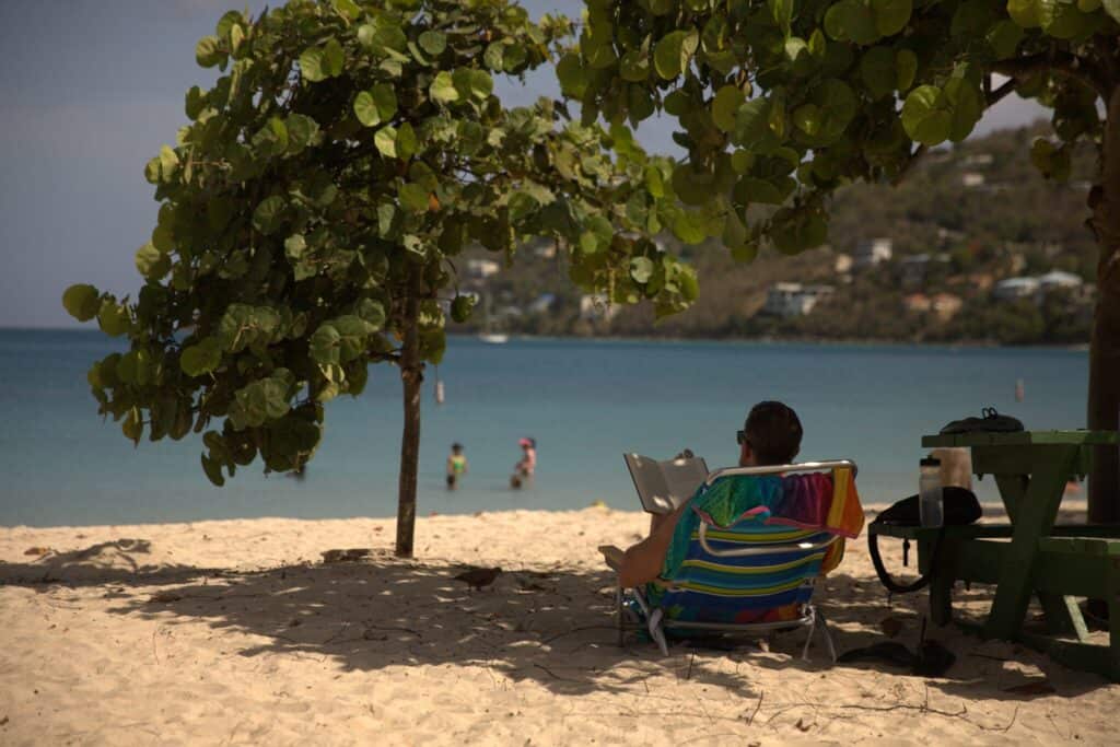 A man sits in a beach chair next to a picnic table under shade while two people wade in the water at Coki Point on St. Thomas in the U.S. Virgin Islands. 