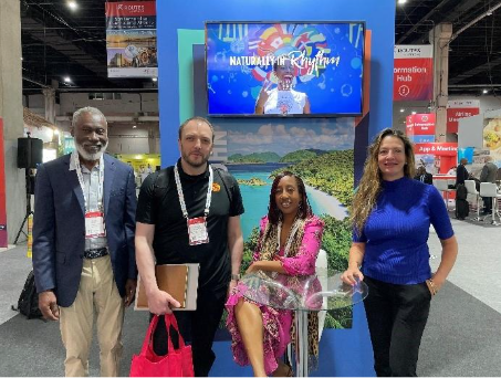 Four people are posing in front of a colorful booth display at a convention or expo, with a screen above them showing the phrase “Naturally in Rhythm.”.