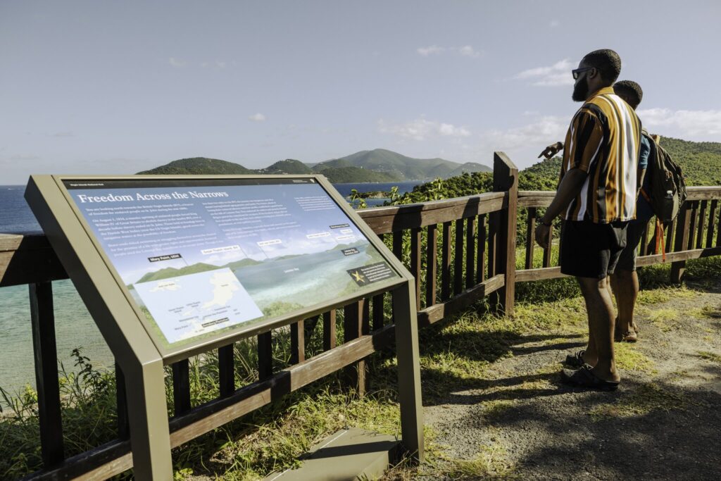 Two people stand by a wooden railing overlooking the ocean and islands, reading an informational sign titled "Freedom Across the Narrows" while touring the ruins of the Annaberg Sugar Plantation on St. John in the USVI.