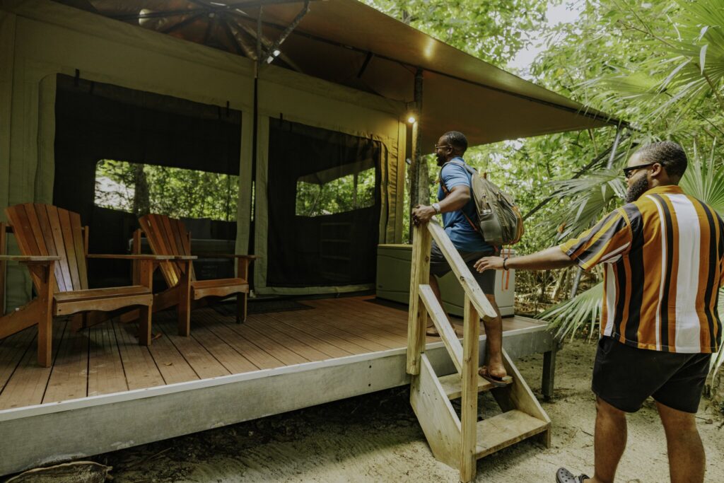 Two people stand on the steps of a tent cabin with wooden chairs on the porch, surrounded by trees and greenery at Cinnamon Bay Beach & Campground on St. John in the USVI.
