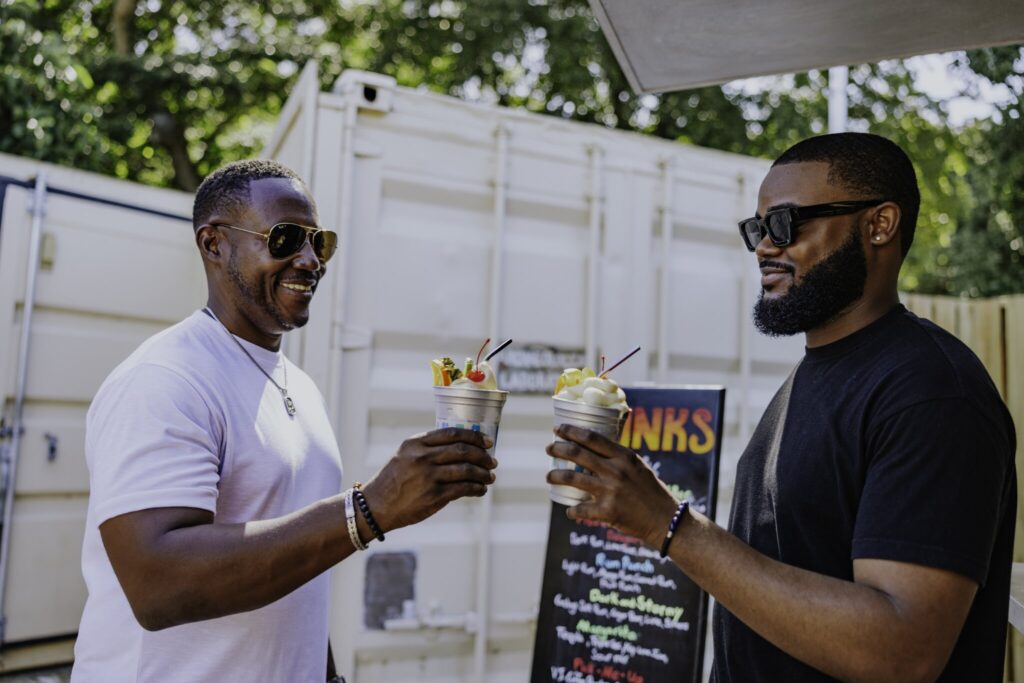 Two men wearing sunglasses clink frozen drinks together outdoors, standing in front of a menu board and a white shipping container at Cinnamon Bay Beach & Campground on St. John in the USVI.