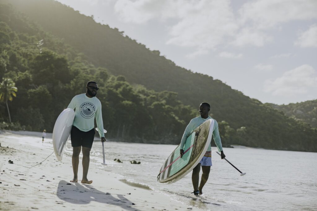 Two people walk on a sandy beach carrying stand-up paddleboards and paddles, with hills and trees in the background.
