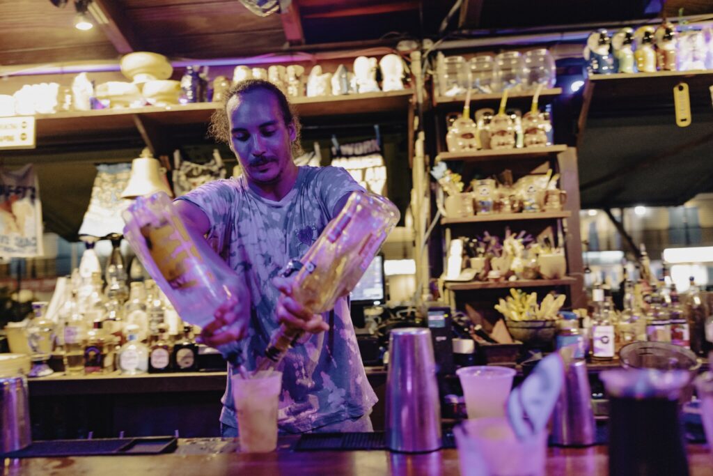 A bartender at Duffy's Love Shack on St. Thomas in the USVI pours liquor from two bottles into a glass at a busy, colorful bar with shelves of bottles and bar tools in the background.