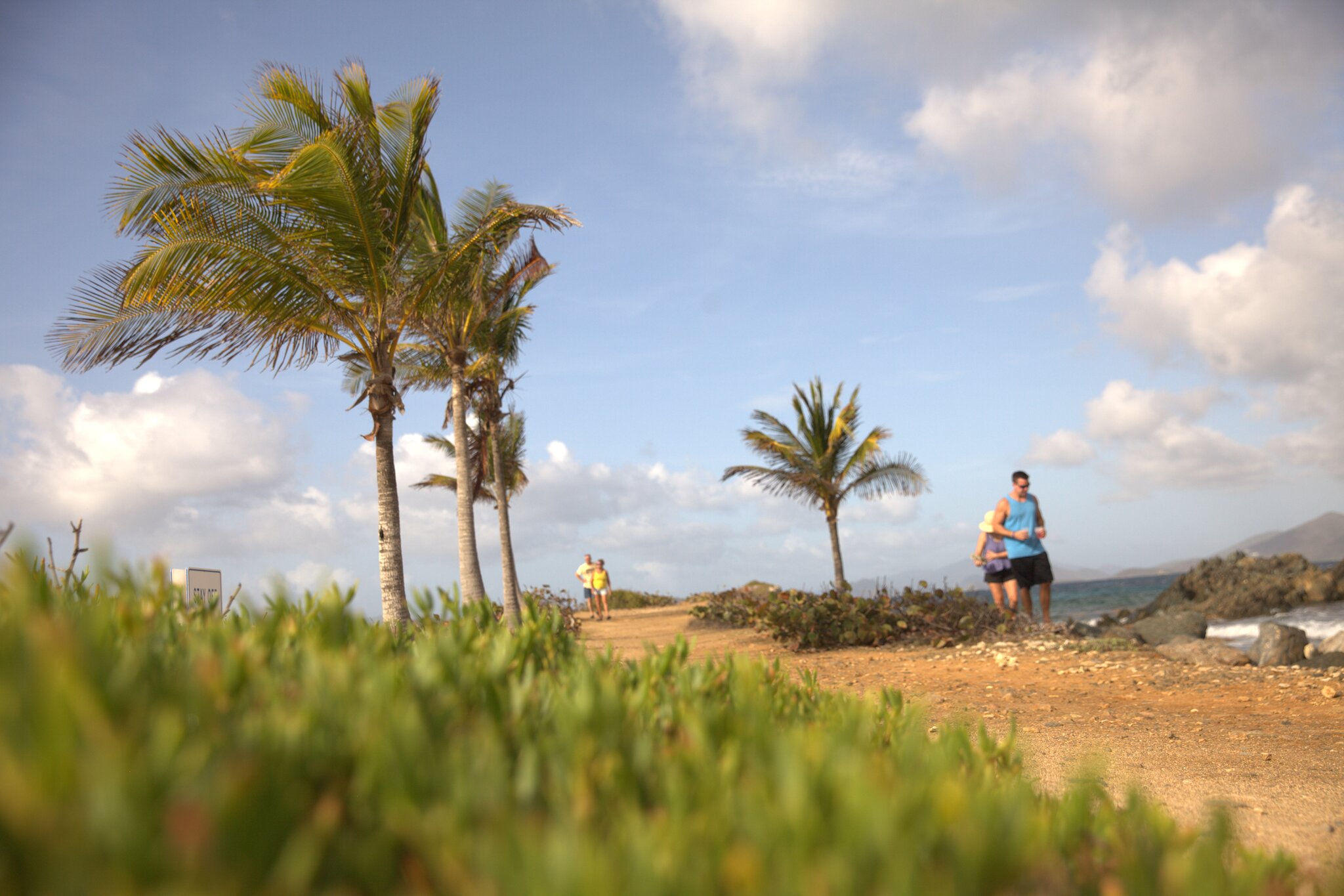 Two people jog on a dirt path by the seaside at Smith Bay Park on St. Thomas, with palm trees and low greenery nearby under a partly cloudy sky.