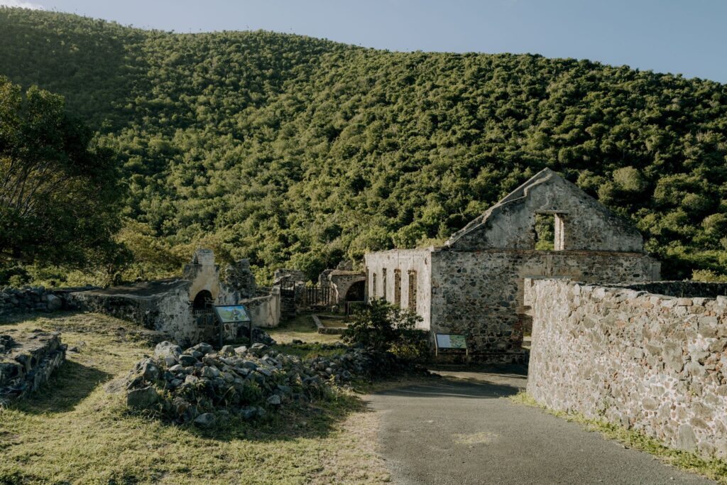 Historic stone ruins ruins of the Annaberg Sugar Plantation on St. John in the USVI stand at the edge of a paved path, surrounded by green hills and trees under a clear sky.