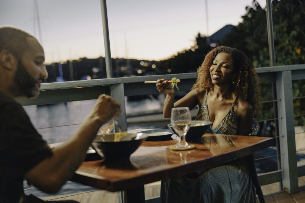 Two people sit at an outdoor restaurant table by the water, eating and drinking, with a sunset and boats visible in the background at Red Hook on St. Thomas in the USVI.