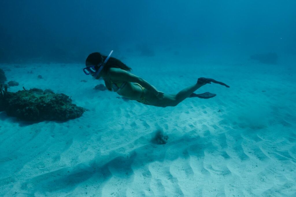A person wearing a snorkel mask and fins swims underwater near the sandy ocean floor with coral formations in the background off the coast of St. Croix in the USVI.
