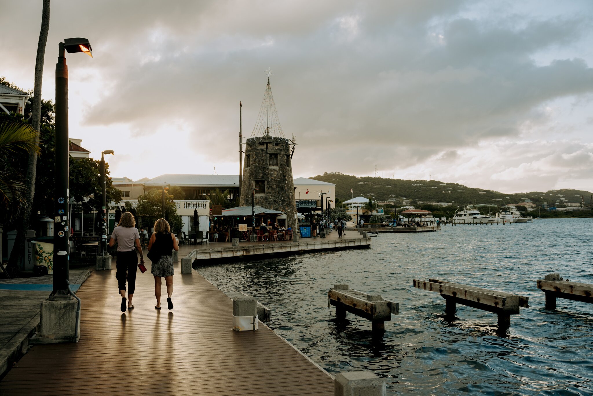Two people walk along the wooden Christiansted Boardwalk by the water at sunset, with buildings, boats and hills in the background in Christiansted on St. Croix in the USVI.