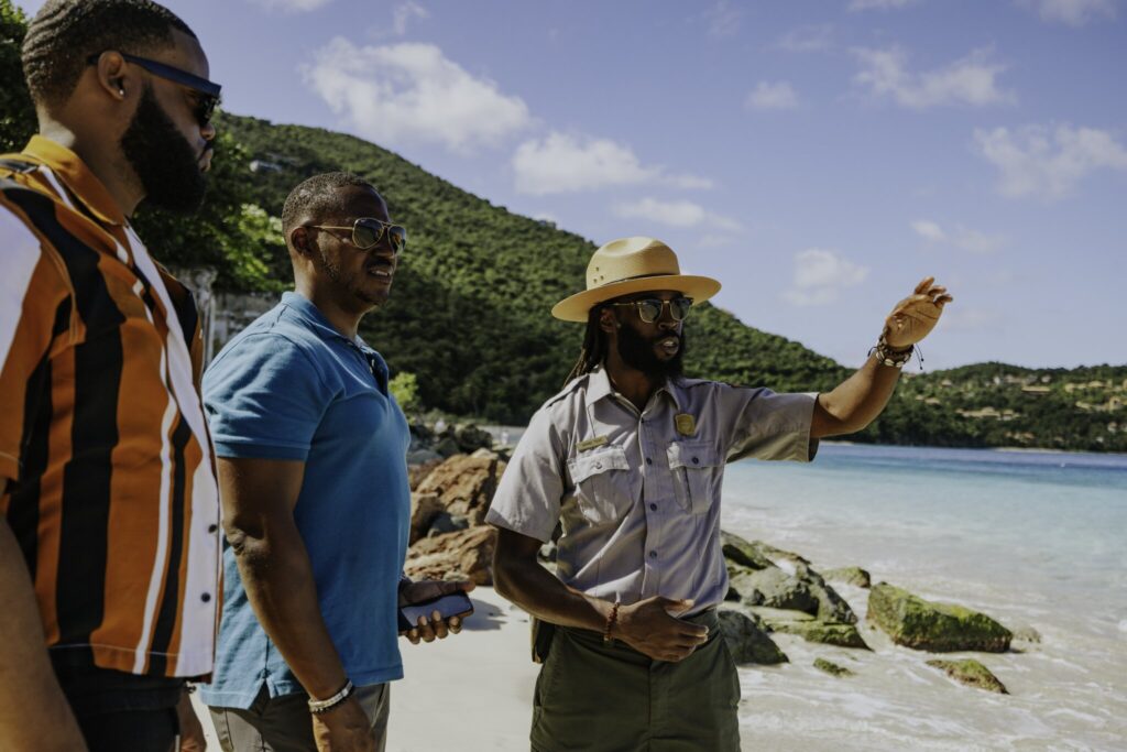 Three men stand on a beach; one, wearing a park ranger uniform and hat, gestures while talking to the other two, who are dressed casually. The ocean and green hills are visible in the background at Cinnamon Bay Beach