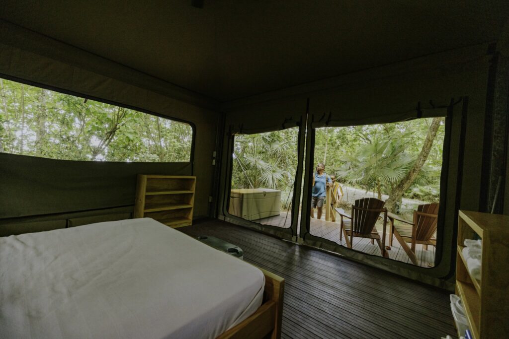Interior of a tented room with a bed and shelves, looking out to a deck with chairs and a person standing outside among trees and greenery at Cinnamon Bay Beach & Campground on St. John, USVI.