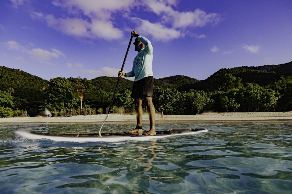 A man stands paddling on a stand-up paddleboard in clear, shallow water near a sandy beach with green hills in the background at Cinnamon Bay Beach on St. John, USVI.