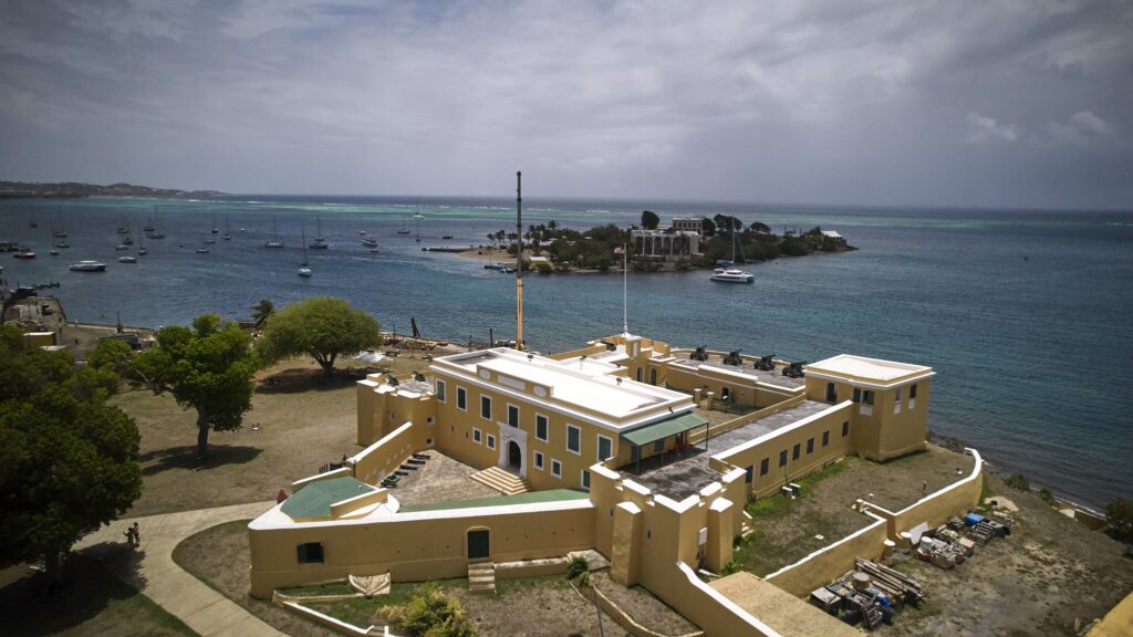 The large, yellow Fort Christiansvaern sits on a coastline overlooking a bay with anchored sailboats and the Protestant Cay island in the distance under a cloudy sky.