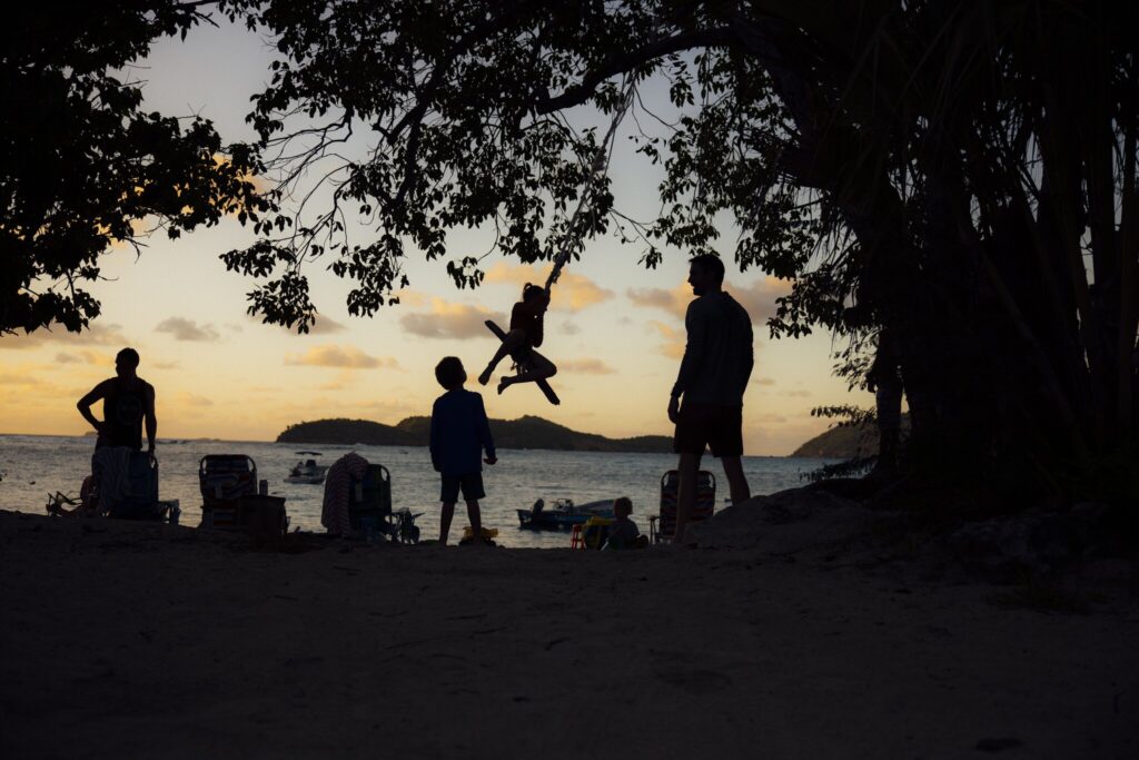 Silhouetted people stand on a beach at sunset; one person is mid-air swinging from a rope tied to a tree, with boats visible on the water in the background at Hideaway at Hull Bay on St. Tghomas, U.S. Virgin Islands