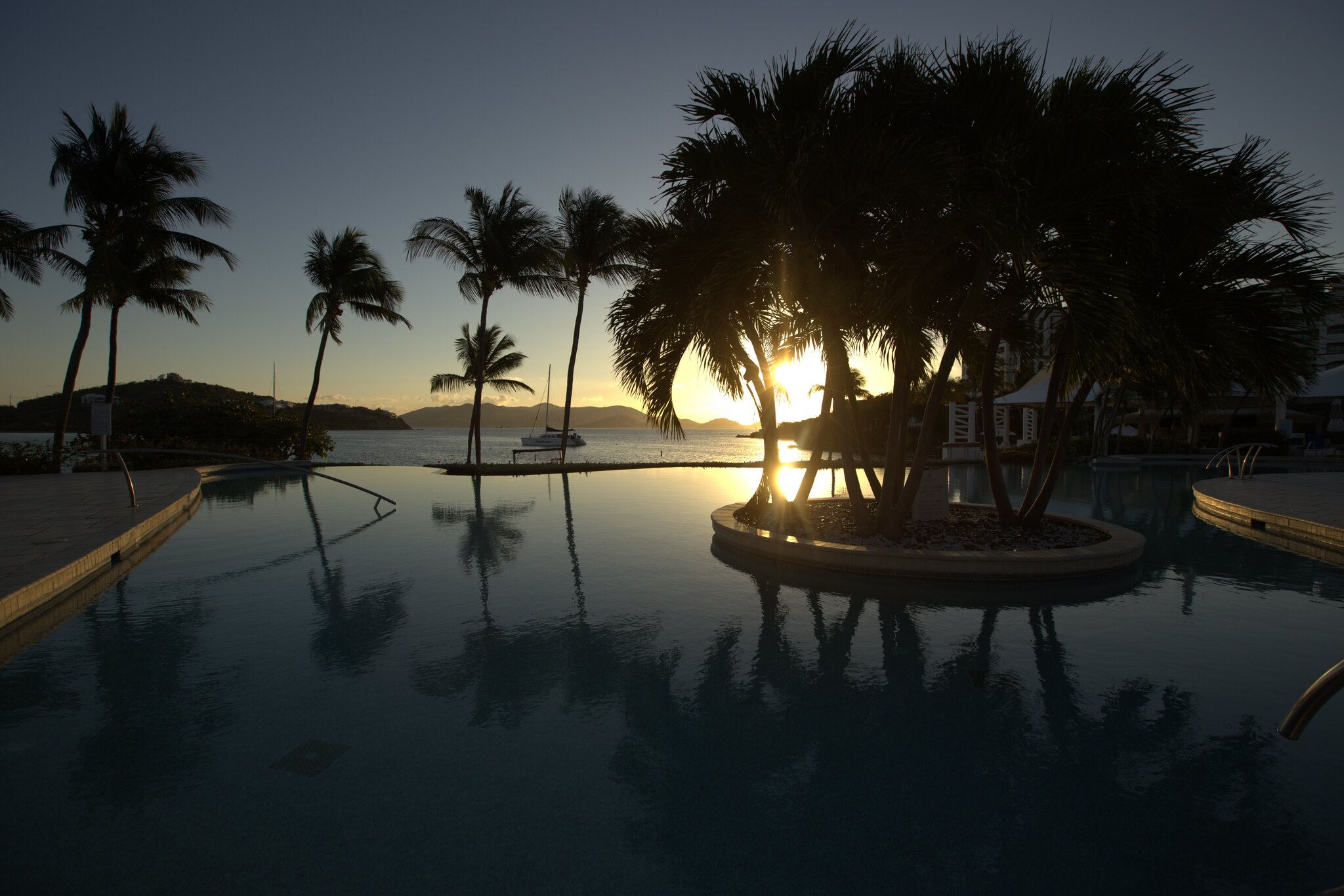 A pool with palm trees at sunset, calm water reflecting the sky, and a boat in the distance.
