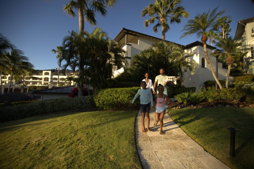 Two adults and two children walk barefoot on a paved path through a landscaped garden, with palm trees and white resort buildings in the background under a clear sky at The Ritz-Carlton, St. Thomas, USVI.