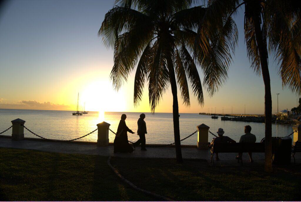 People walk and sit by the waterfront at sunset with the Frederiksted Pier in view and palm trees, a calm sea, and sailboats in the background on St. Croix, USVI.
