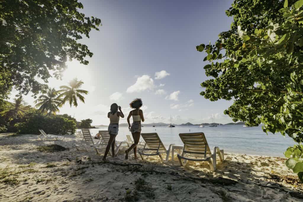 Two people walk along a sandy beach lined with lounge chairs, surrounded by tropical trees, with boats visible on the water under a clear sky at Honeymoon Beach on St. John, USVI.