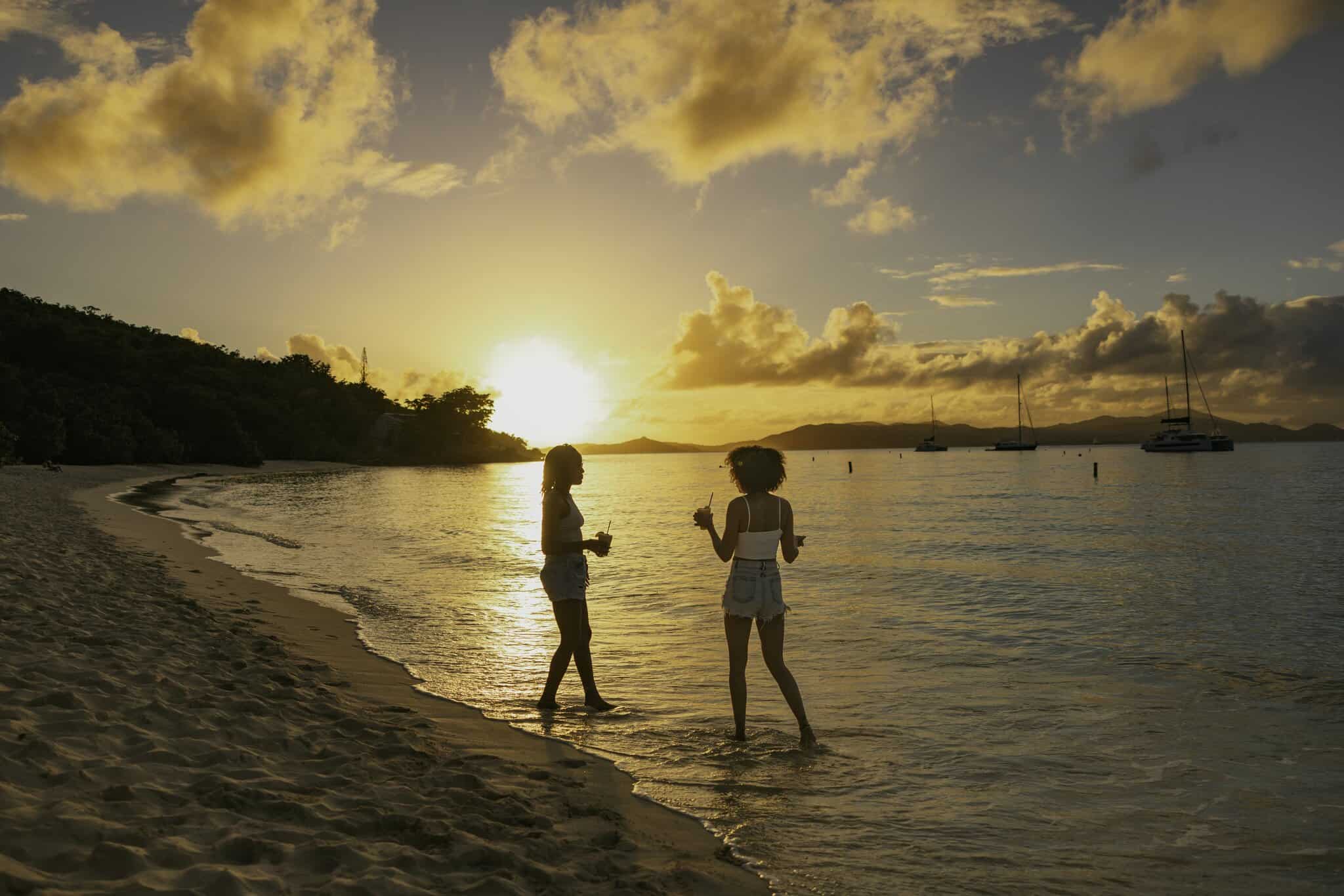 Two people stand at the edge of the water on a sandy beach at sunset, with boats anchored in the background and clouds in the sky at Honeymoon Beach on St. Jolhn, USVI.