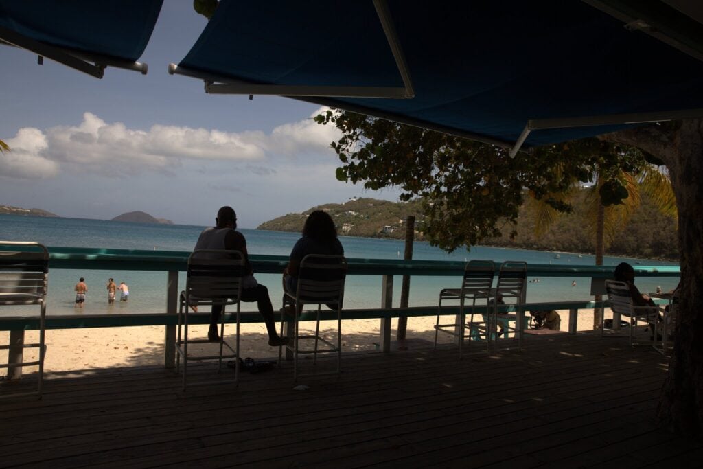 Two people sit on chairs under shade, facing a beach and calm water, while others stand in the shallow sea under a partly cloudy sky at Coki Point Beach, St. Thomas, USVI..