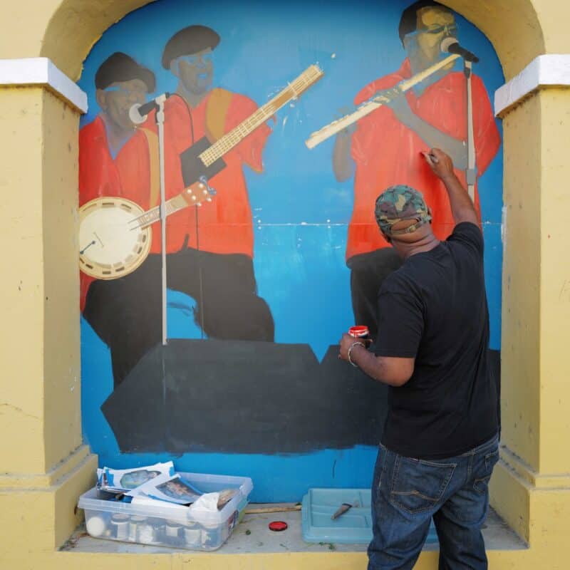 An artist paints a mural of Quelbe musicians in red shirts on a blue background while standing between two yellow columns of a building on St. Croix, USVI. Art supplies are on the ground nearby.