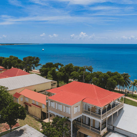Aerial view of buildings with red roofs near a coastline, overlooking blue ocean water and a partly cloudy sky.