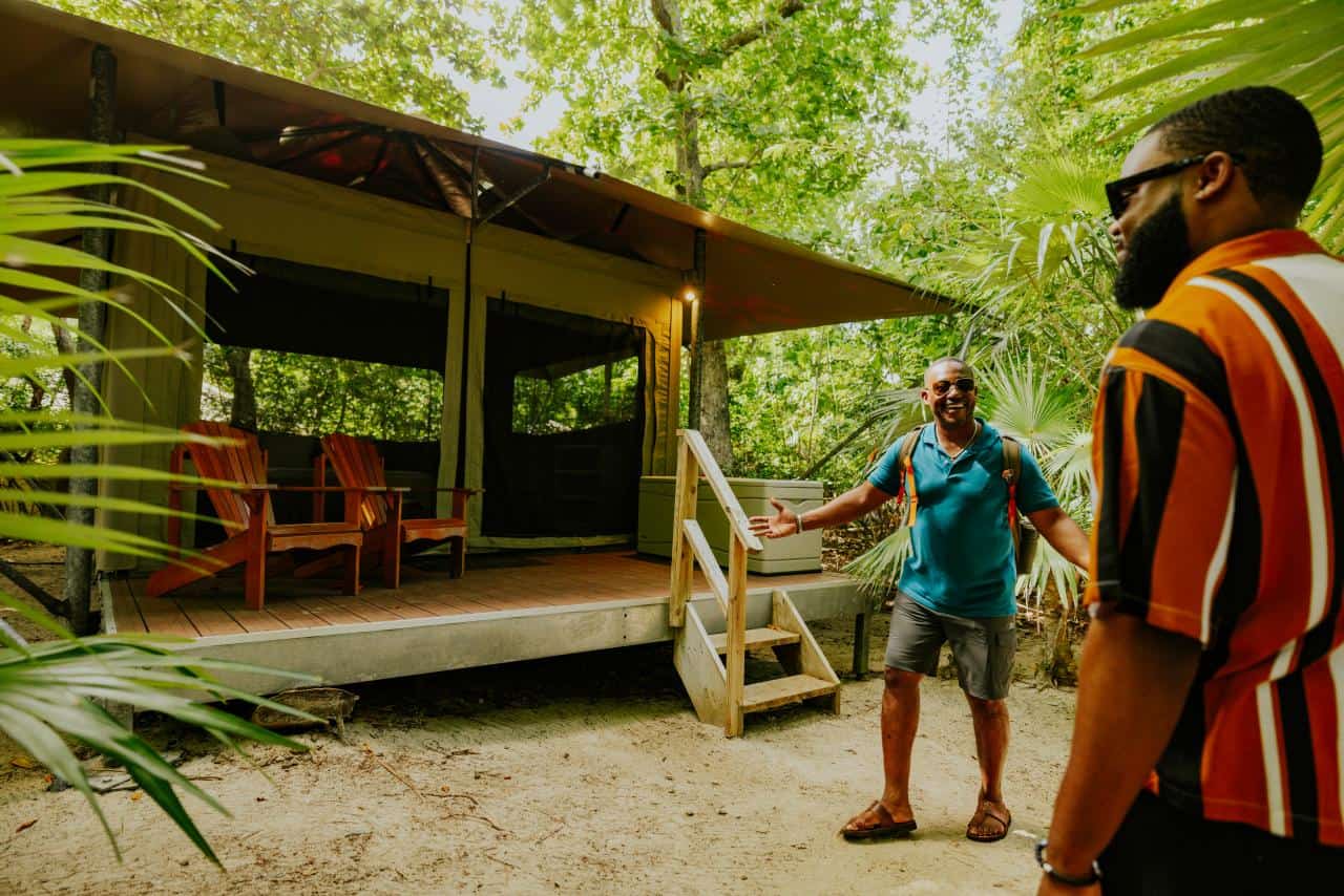 Two men stand outside a canvas glamping tent surrounded by trees and plants, with wooden chairs on the porch t Cinnamon Bay Beach & Campground on St. John in the USVI.