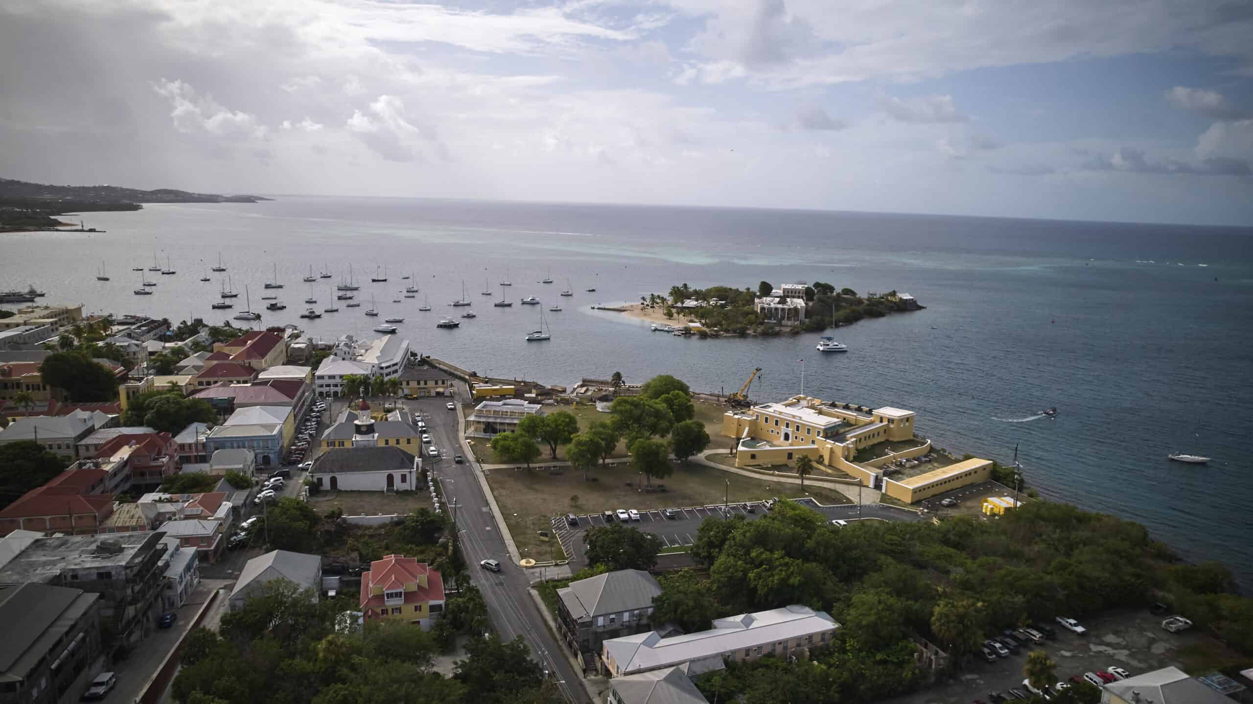 Aerial view of the coast of Christiansted on St. Croix, U.S. Virgin Islands, with colorful buildings, a harbor filled with boats, and a small island near the shore under a partly cloudy sky.
