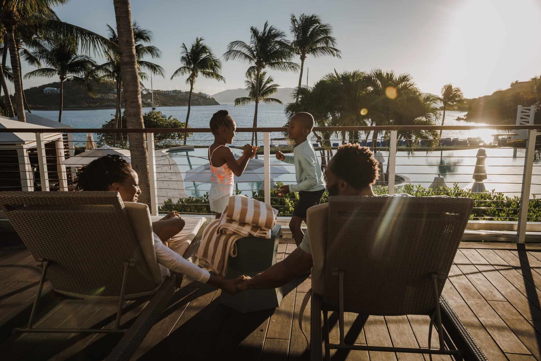 Two adults relax on lounge chairs while two children stand nearby on a deck at The Ritz-Carlton, St. Thomas overlooking palm trees, water, and a sunset.