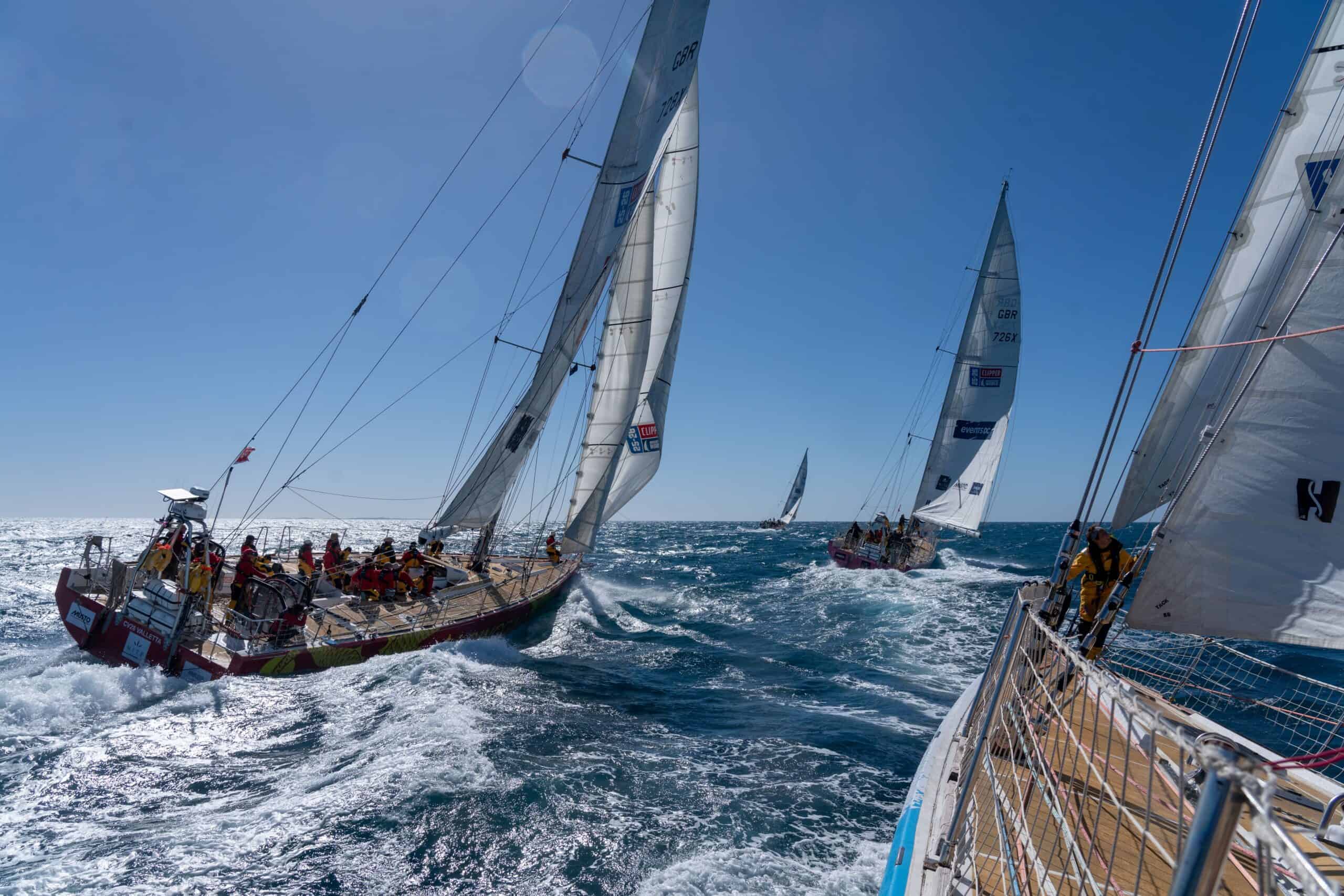 Several sailboats with crews race on choppy blue water under a clear sky, their sails fully extended as they navigate through the waves.