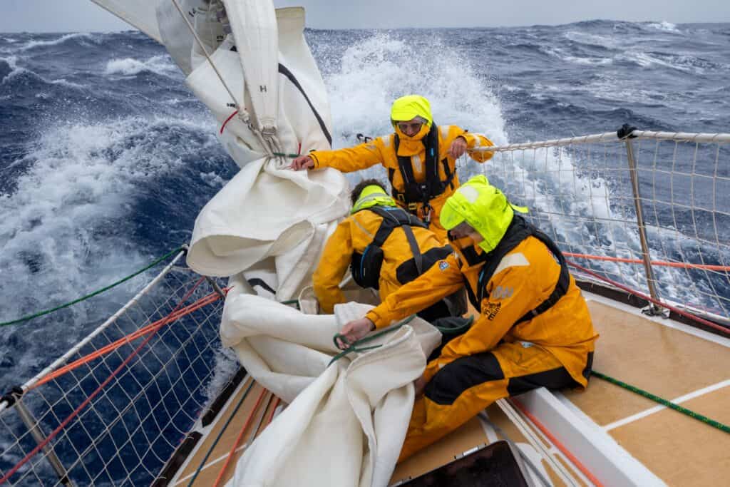 Three sailors in yellow rain gear secure sails on a Clipper Race sailing yacht in rough seas, with waves crashing over the side and cloudy skies overhead.