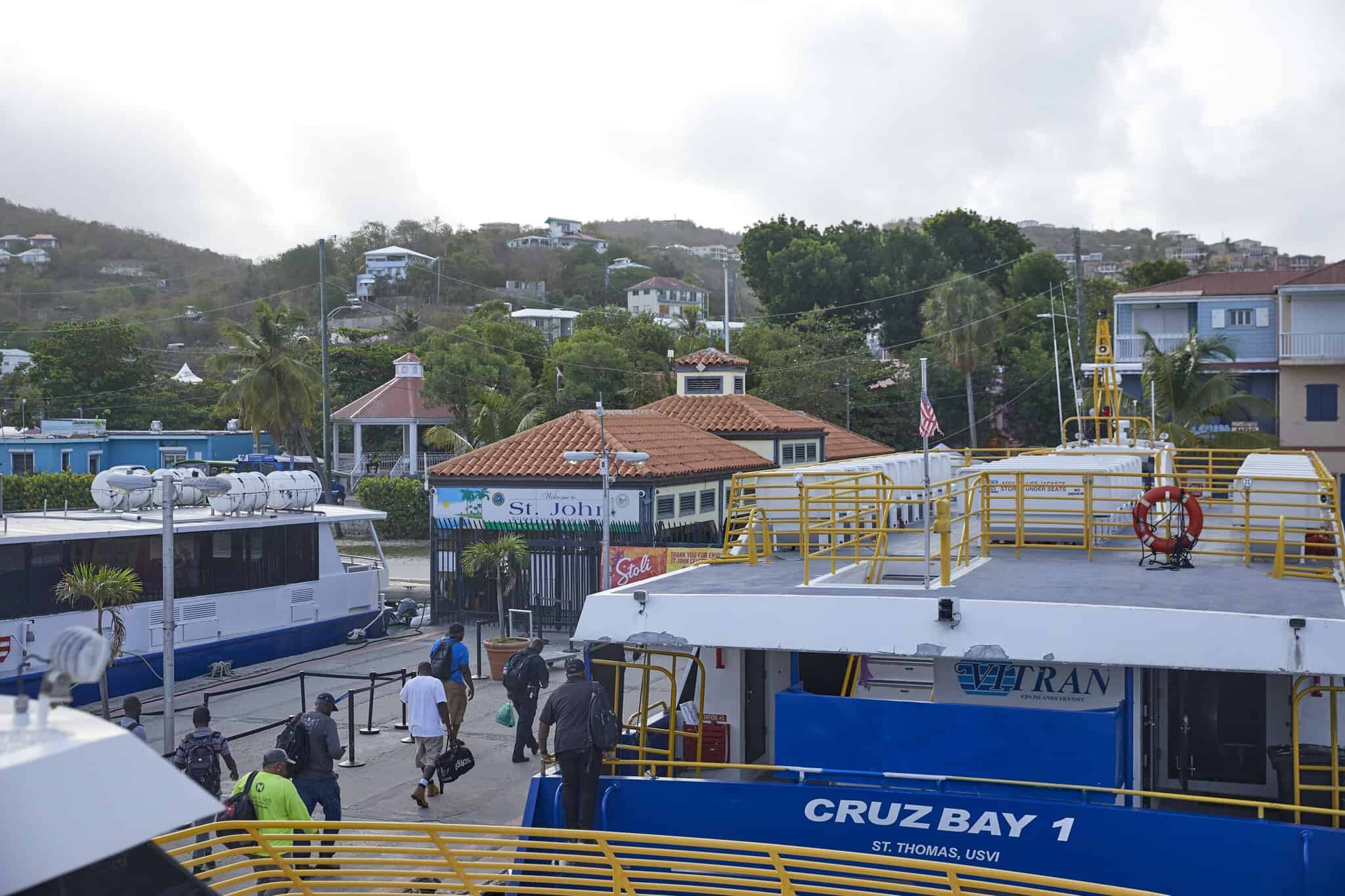 People disembark from ferries at a dock in Cruz Bay, St. John, US Virgin Islands, with houses and trees visible on the hillside in the background.