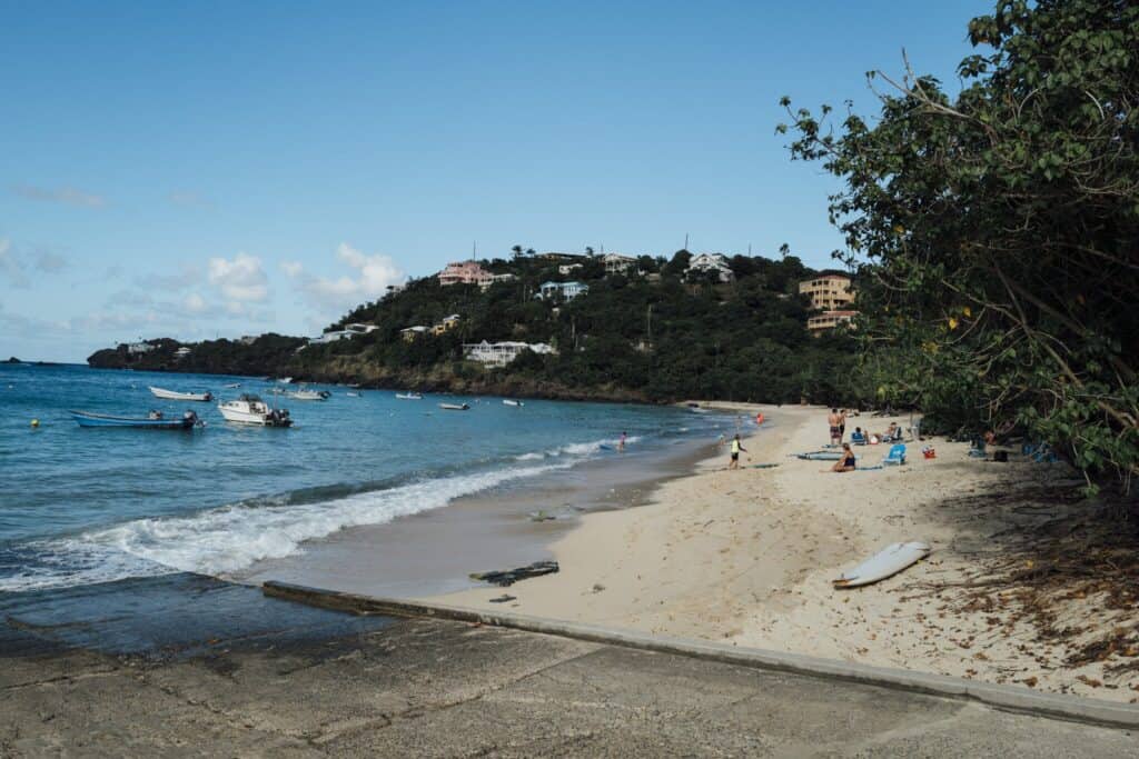 A sandy beach with people relaxing near the water, several small boats anchored offshore, and a tree-lined hillside with houses in the background under a clear blue sky at The Hideaway at Hull Bay on St. Thomas, USVI.