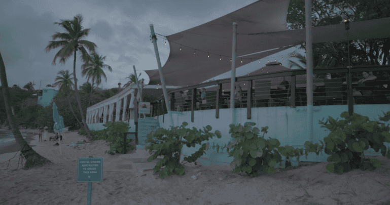 Outdoor beachfront restaurant at Secret Harbor Beach Resort on St. Thomas, USVI, with string lights and patrons seated under canopies. Palm trees, beach and a posted beach sign visible in the foreground. Evening sky overhead.
