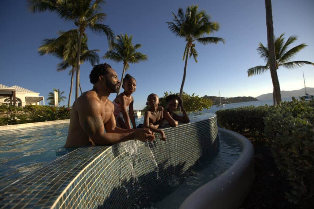 A man. woman and two children lean on the edge of a pool surrounded by palm trees, with clear skies and water in the background at The Ritz-Carlton, St. Thomas, USVI.